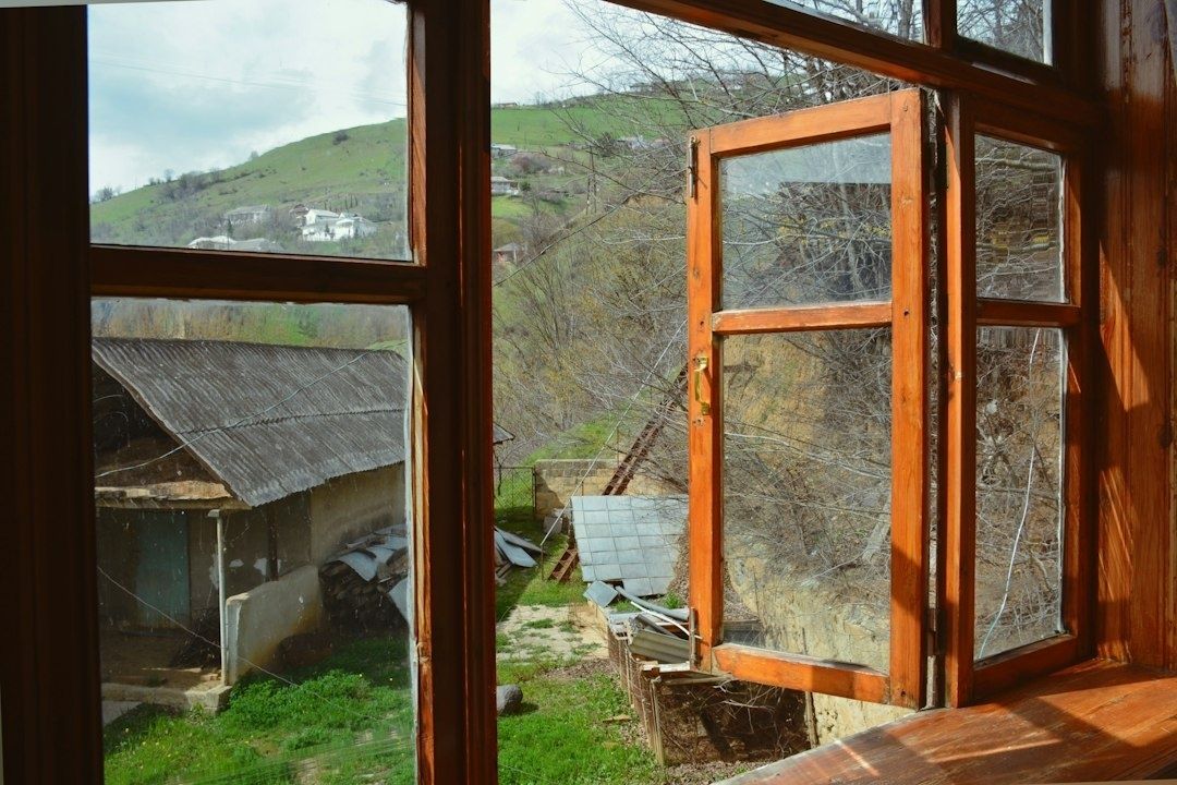 Geöffnetes Holzfenster mit Blick auf grüne Hügel und ein Landhausgrundstück